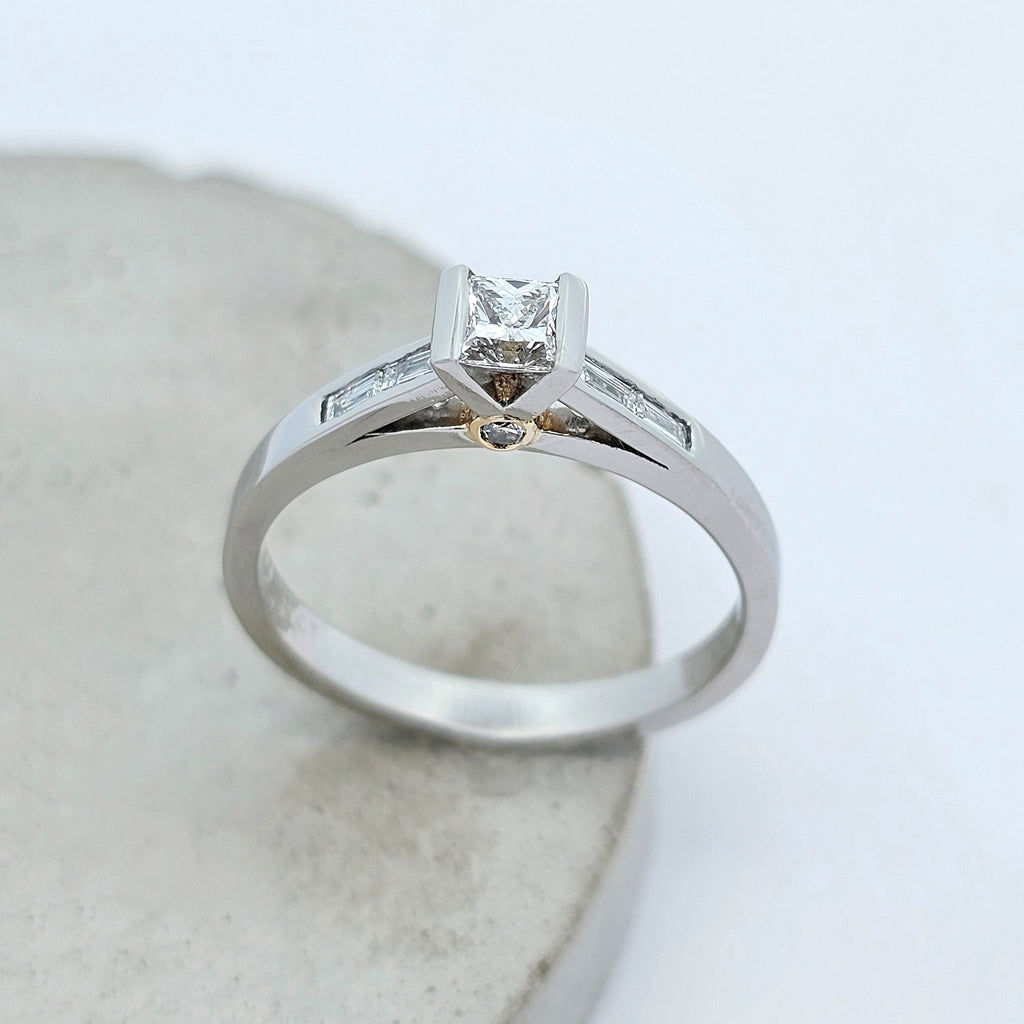 Platinum ladies engagement ring featuring an elevated semirub set princess cut diamond, stepping down to shoulders channels set with baguette diamonds. There is a little round white diamond hidden under the setting on each side in a touch of yellow gold.