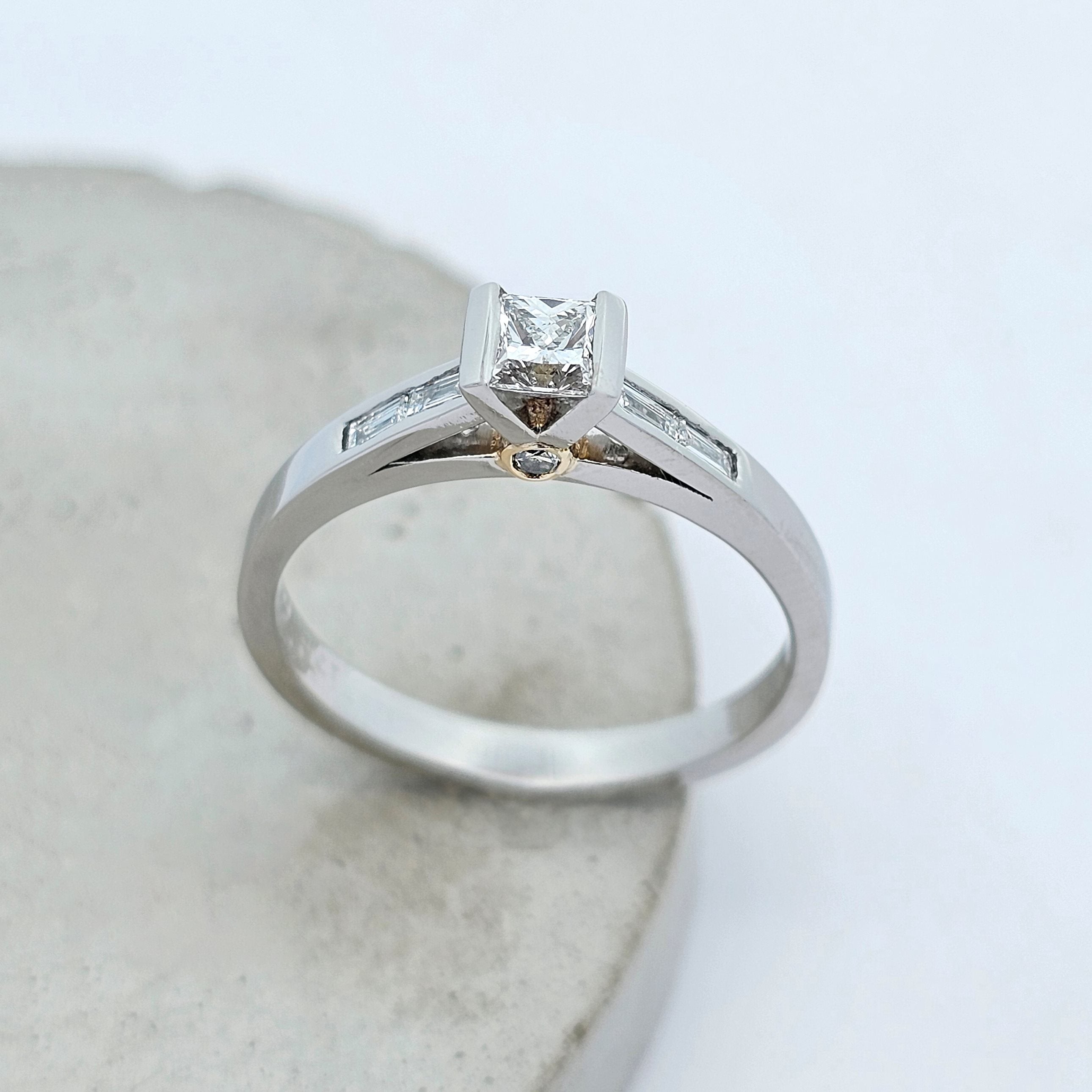 Platinum ladies engagement ring featuring an elevated semirub set princess cut diamond, stepping down to shoulders channels set with baguette diamonds. There is a little round white diamond hidden under the setting on each side in a touch of yellow gold.