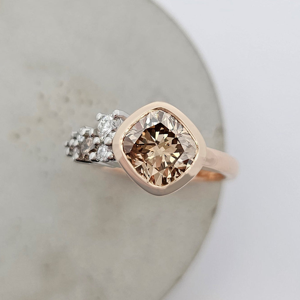 18ct rose gold and platinum ring featuring a large cushion shaped champagne diamond and asymmetrical claw set cluster of small round white diamonds.