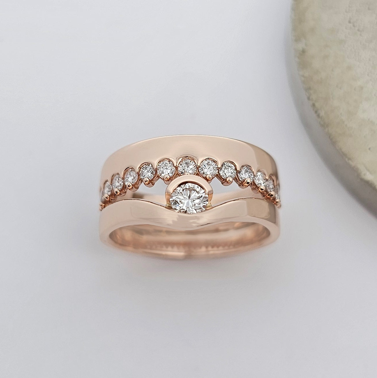 9ct rose gold ring with a row of small claw set white diamonds in a wide band of solid gold opposite a curved band that sweeps around a solitaire diamond.