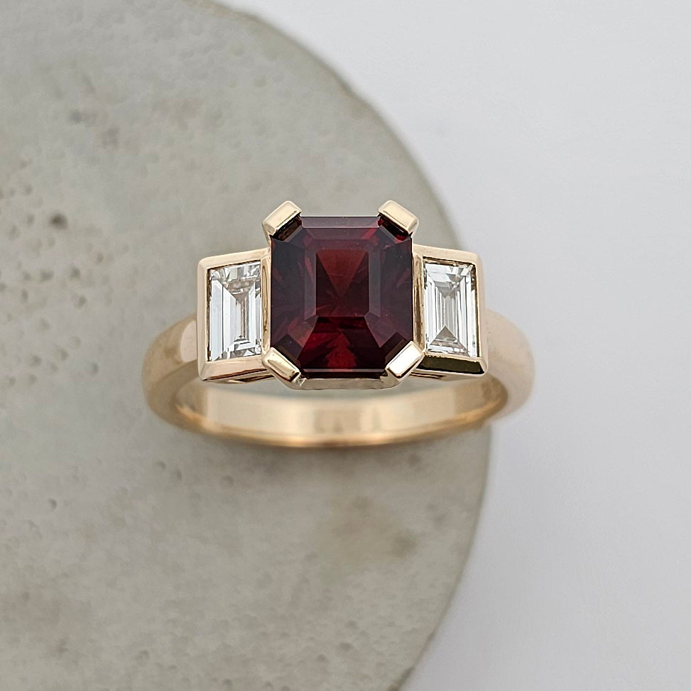 9ct yellow gold ladies ring with four claw setting holding a square emerald cut garnet between rubover set white baguette diamonds with a wide gold band.