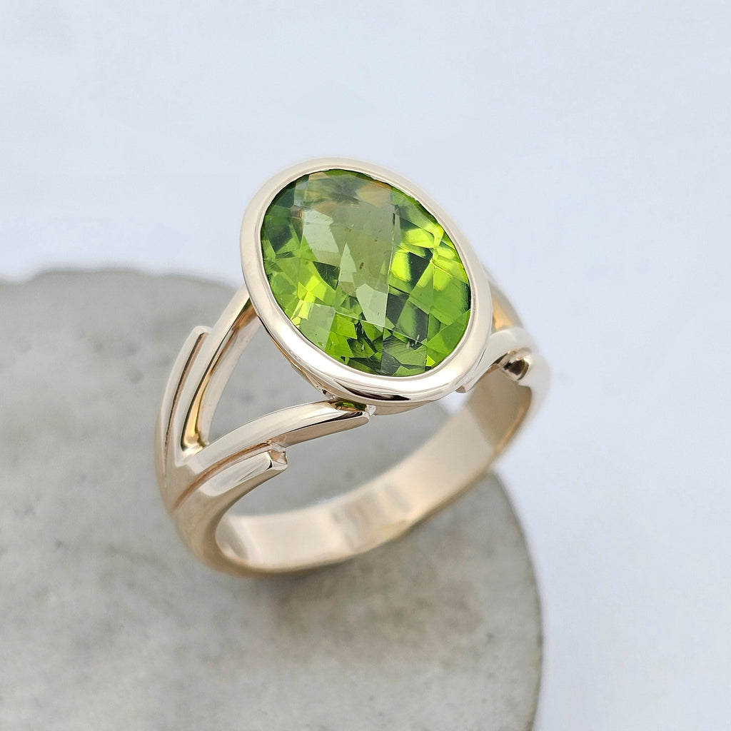 Enchanted oval checkerboard cut peridot ring in yellow gold with rub-over setting and split shoulders.