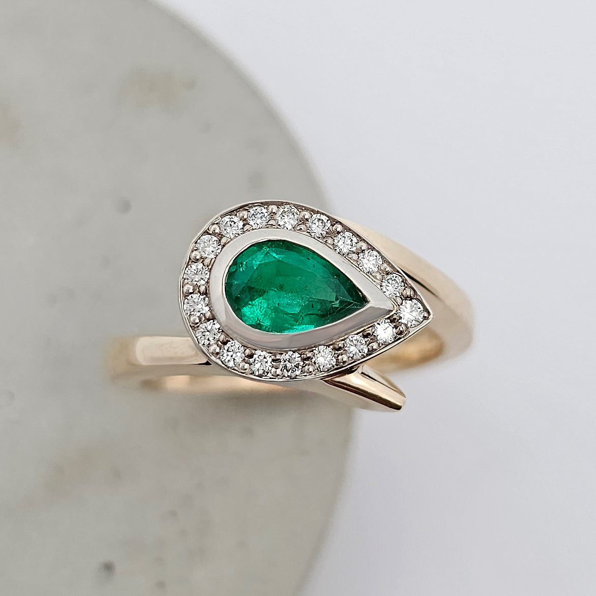9ct yellow and white gold ladies ring with a rub over set pear shaped green emerald surrounded by a halo of bead set white diamonds in a cross over style band.