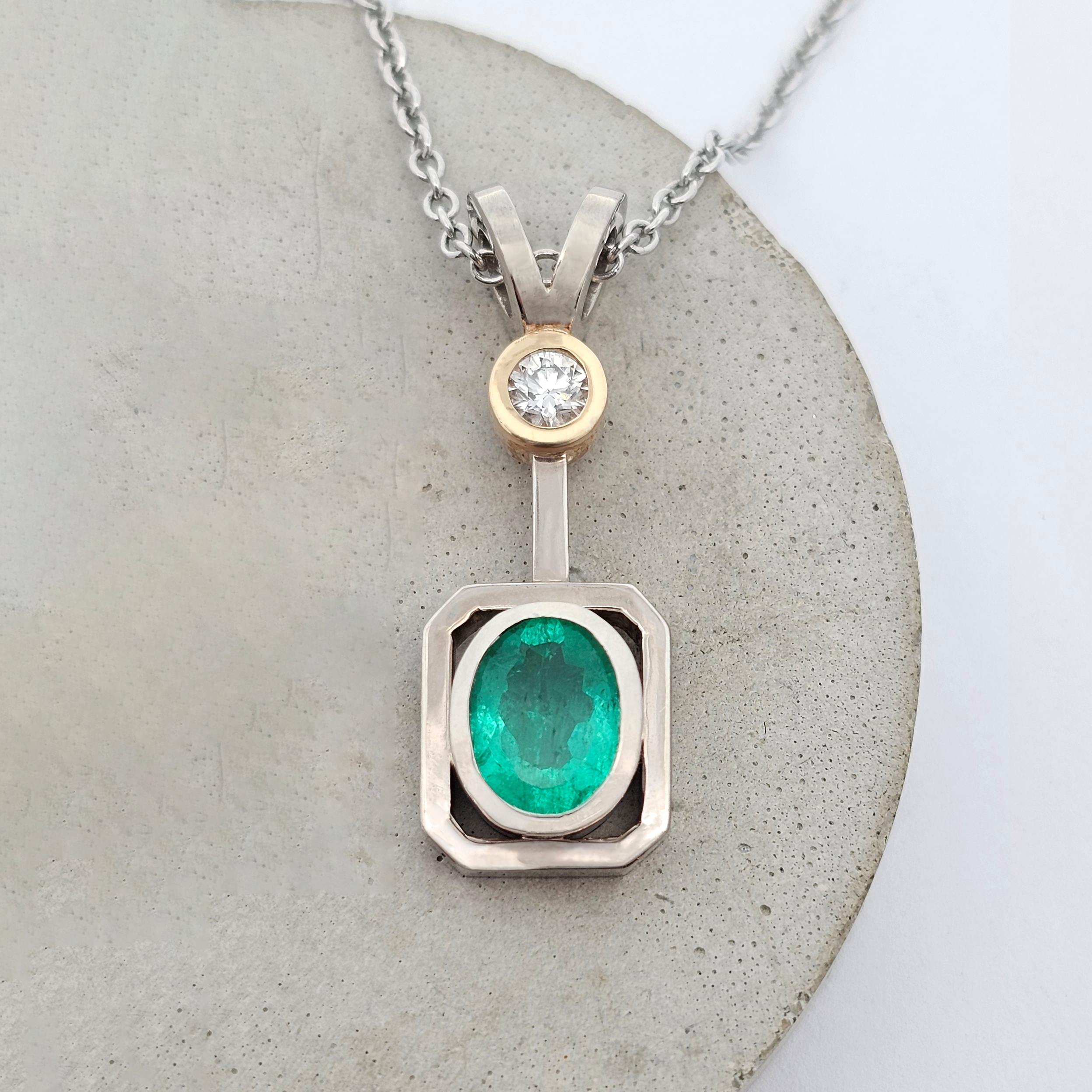 White gold pendant featuring an oval emerald in a geometric frame with a yellow gold bezel-set diamond above.