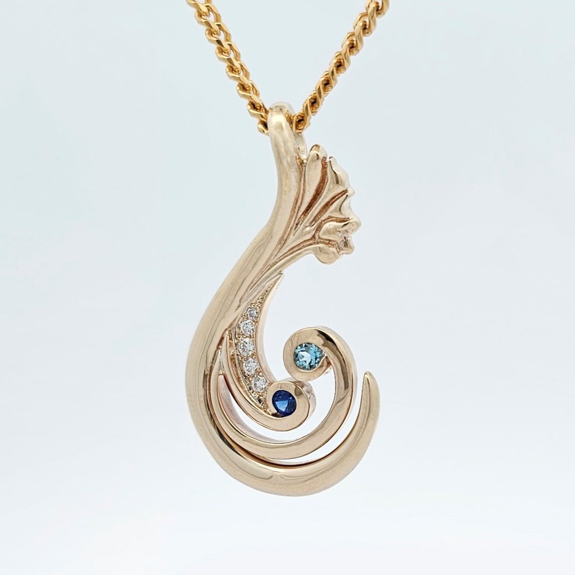 9ct yellow gold organically carved, textural pendant featuring two koru wrapping around a deep blue sapphire and clear blue aquamarine, with a line of round white diamonds.