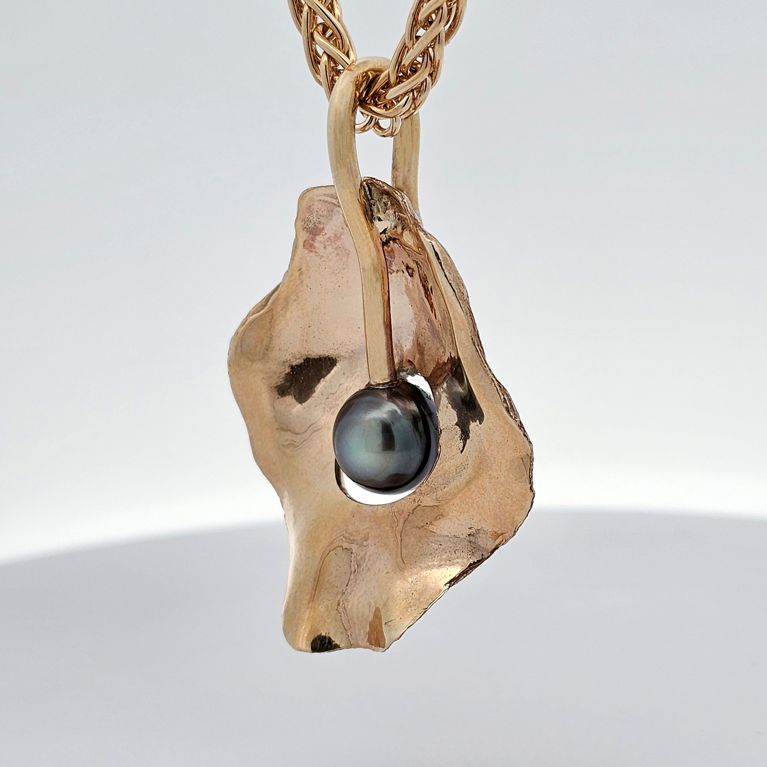 Reverse side of the Golden Oyster pendant in solid yellow gold, showing textured, natural oyster-shell details with a glowing grey-black pearl set in the centre, suspended from a woven gold chain