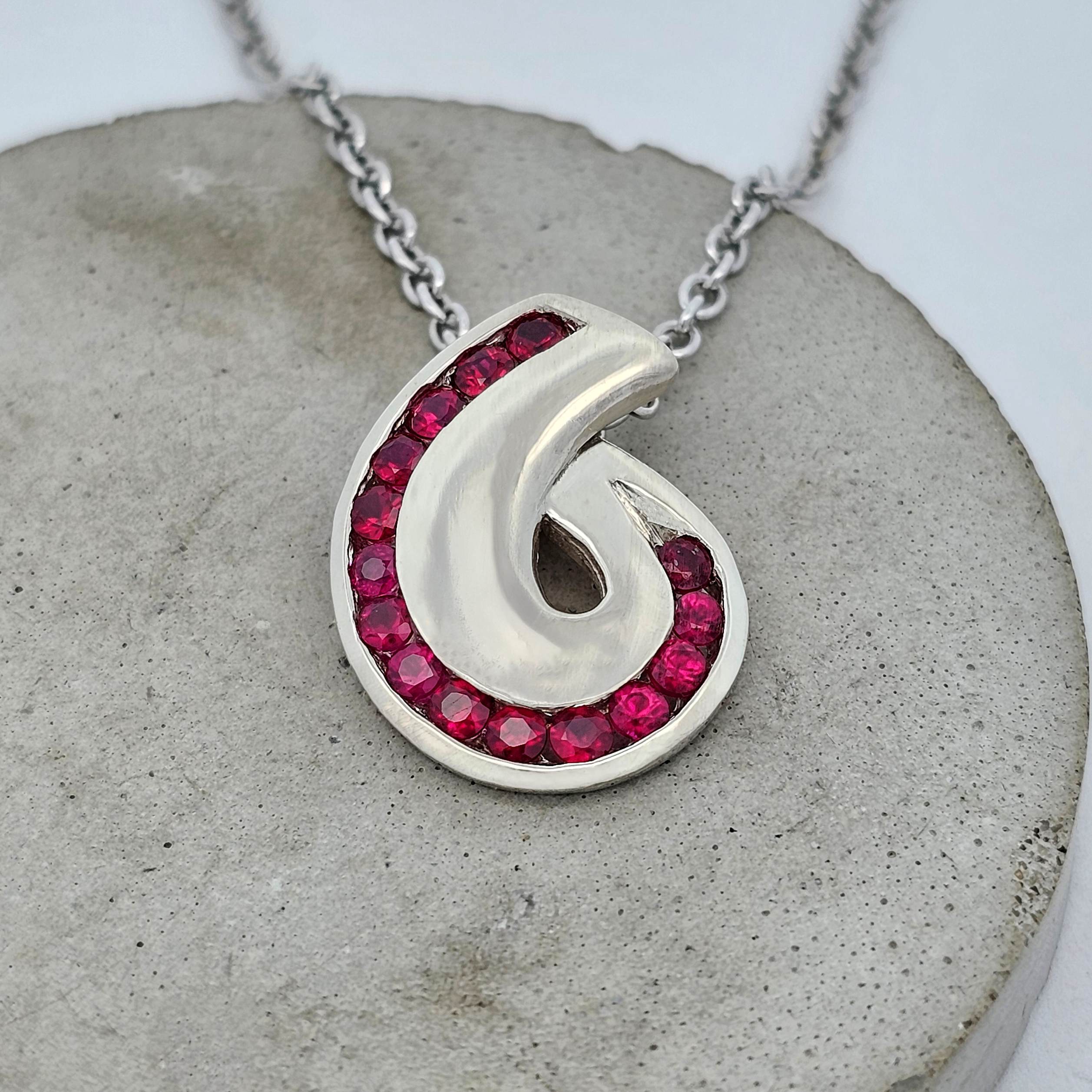 Koru-inspired ruby pendant featuring a spiral design set with vivid red round rubies in polished metal, creating a bold and flowing form.